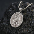 Load image into Gallery viewer, Polished silver Saint Roch pendant necklace on textured volcanic rock, centered macro jewelry photo with detailed Catholic medal engraving and bright silver highlights.
