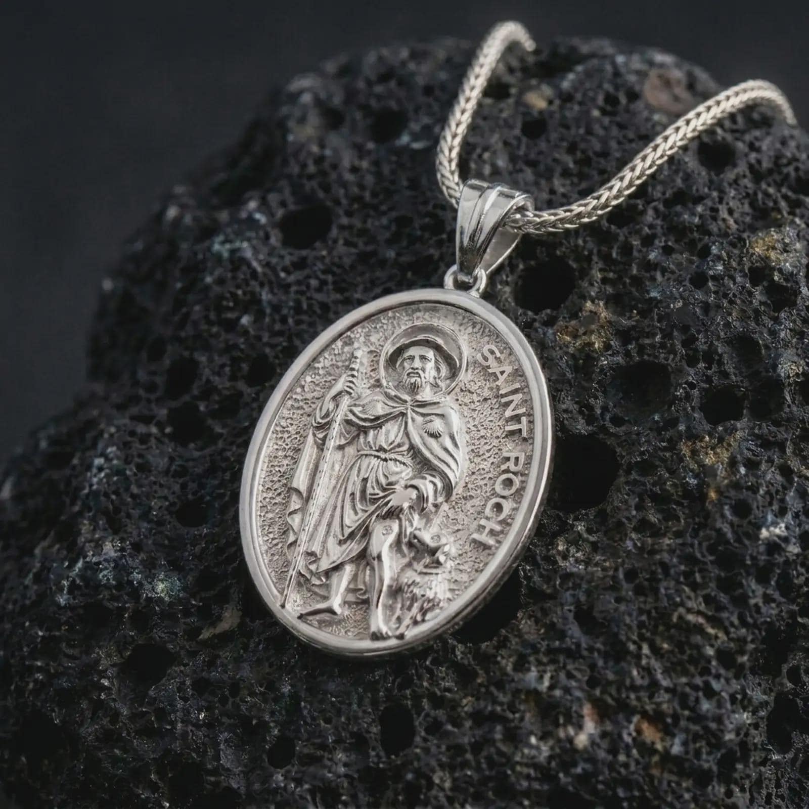 Polished silver Saint Roch pendant necklace on textured volcanic rock, centered macro jewelry photo with detailed Catholic medal engraving and bright silver highlights.