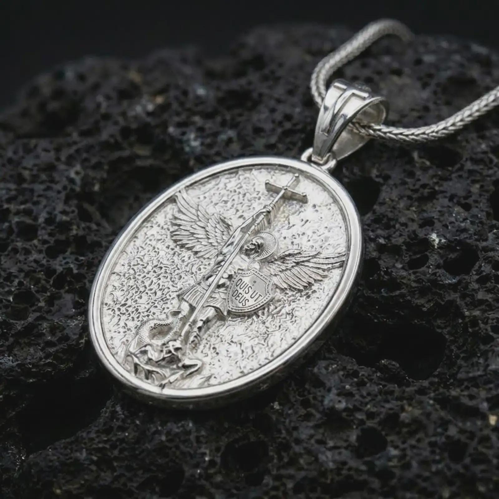 Polished silver Saint Michael pendant necklace on textured volcanic rock, centered macro jewelry photo with detailed archangel relief and bright silver highlights.