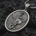 Load image into Gallery viewer, Oxidized Saint Michael pendant necklace on textured volcanic rock, close-up studio shot highlighting the sword, wings, shield, fallen demon detail, and antique silver contrast.
