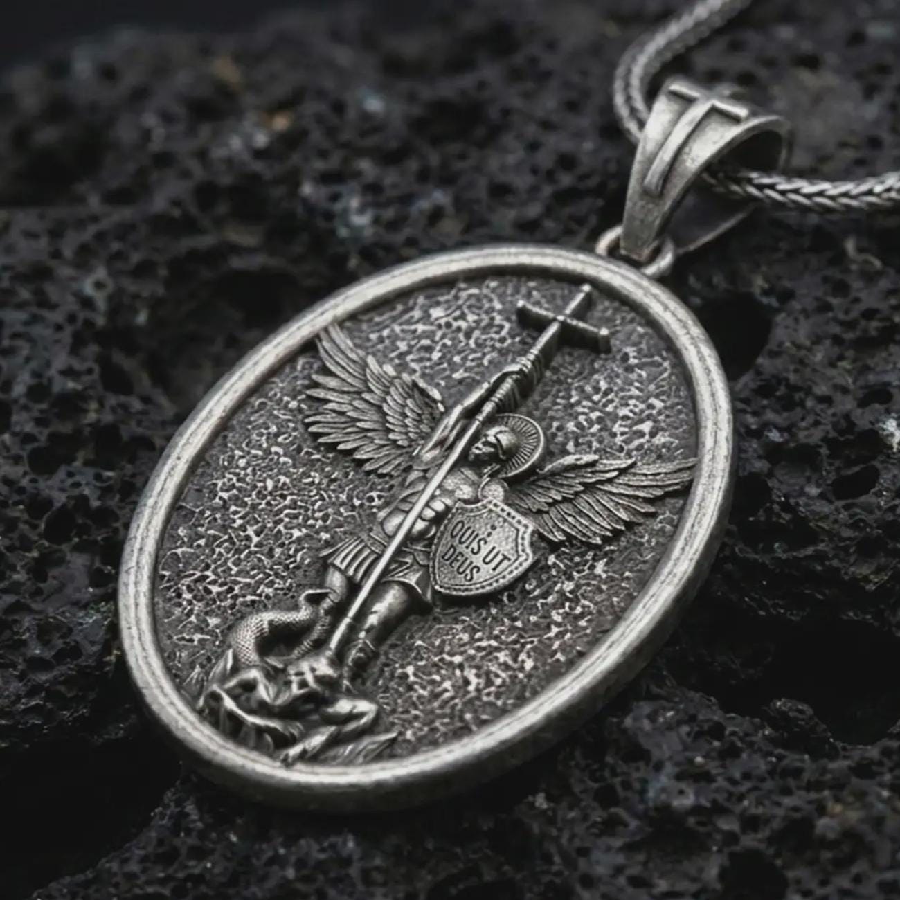 Oxidized Saint Michael pendant necklace on textured volcanic rock, close-up studio shot highlighting the sword, wings, shield, fallen demon detail, and antique silver contrast.