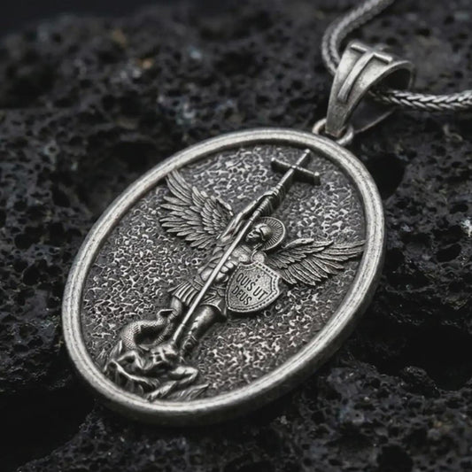 Oxidized Saint Michael pendant necklace on textured volcanic rock, close-up studio shot highlighting the sword, wings, shield, fallen demon detail, and antique silver contrast.