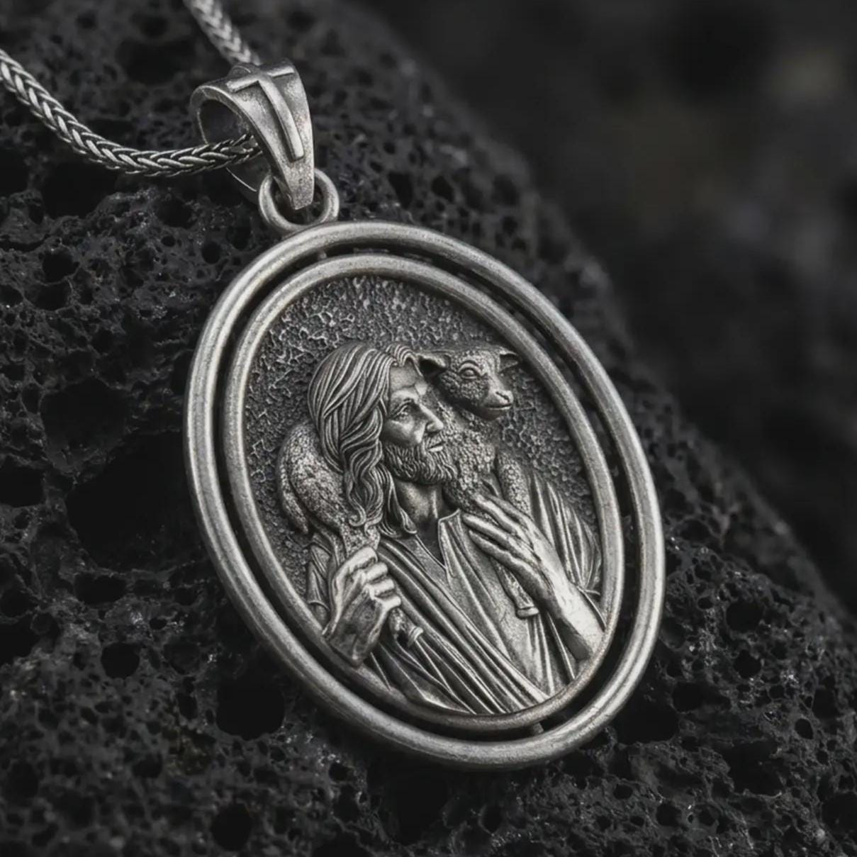 Oxidized spinning Good Shepherd pendant necklace on textured volcanic rock, close-up studio shot highlighting the Jesus and lamb relief with antique silver contrast.