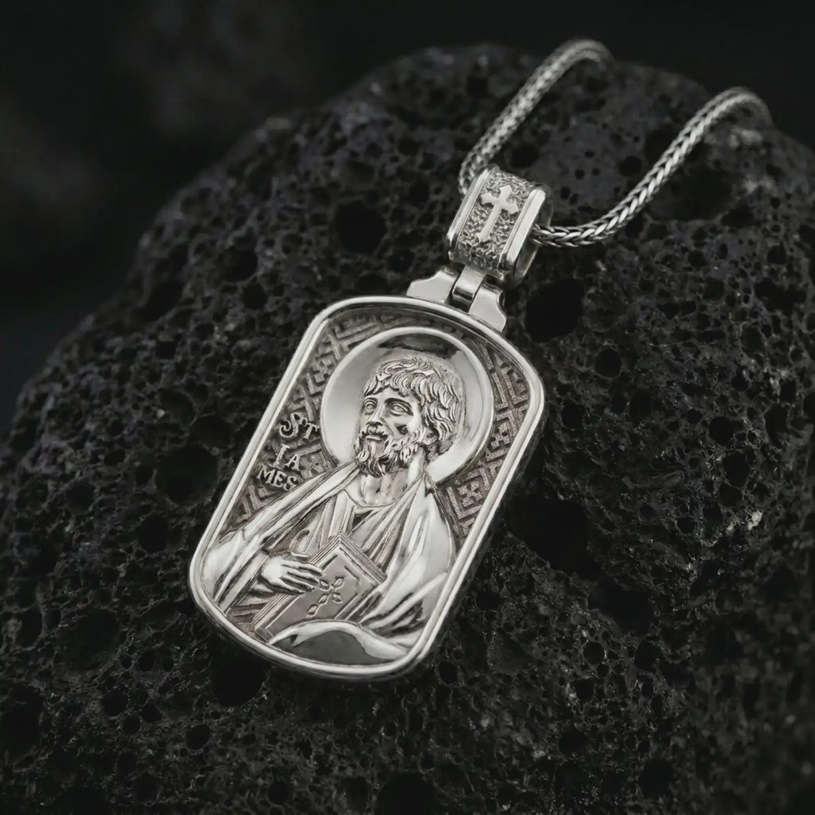 Polished silver Saint James the Apostle pendant necklace on textured volcanic rock, centered macro jewelry photo with detailed Catholic medal engraving and bright silver highlights.