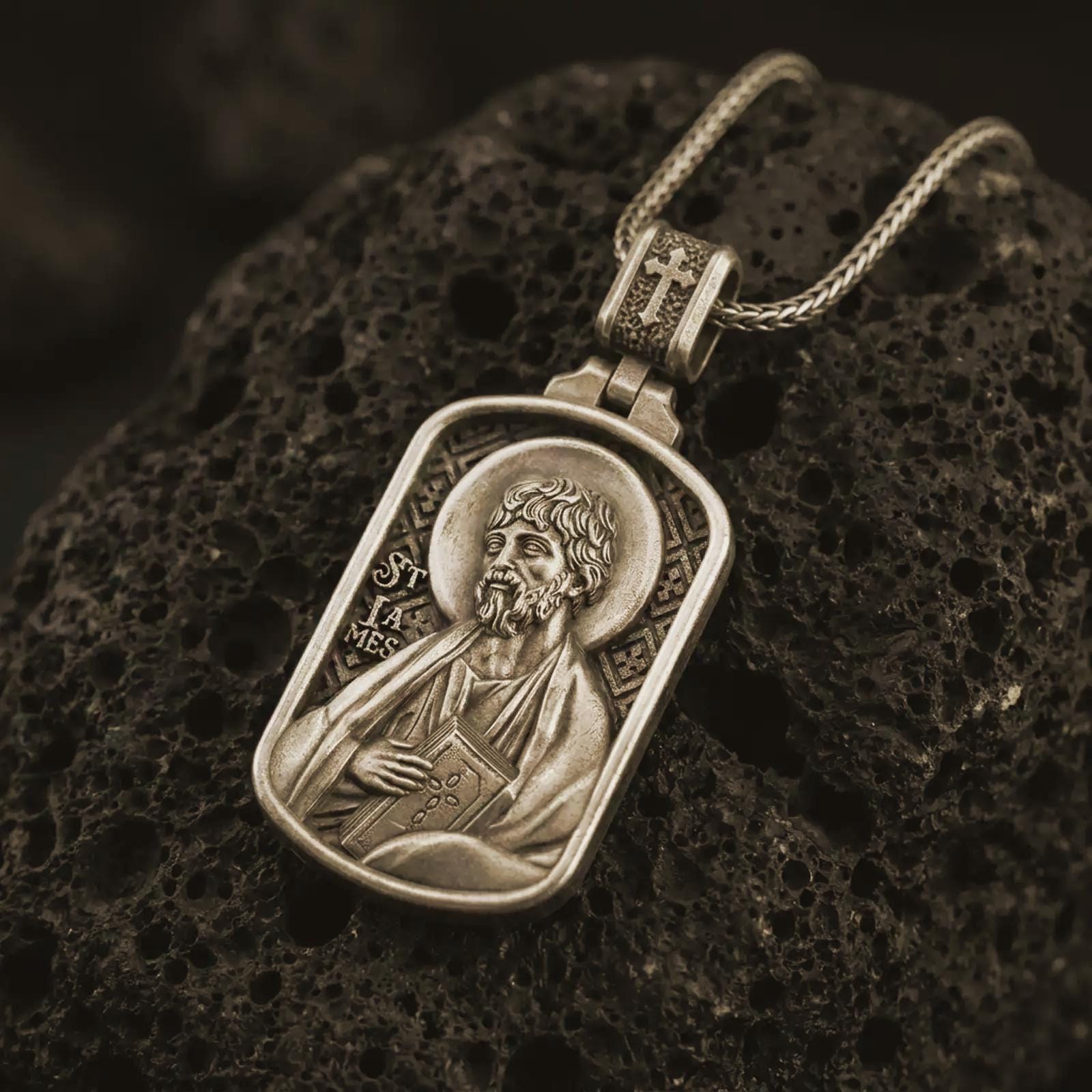 Bronze Saint James the Apostle pendant necklace on textured volcanic rock, close-up studio shot highlighting the apostle relief, halo, book detail, and antique silver contrast.