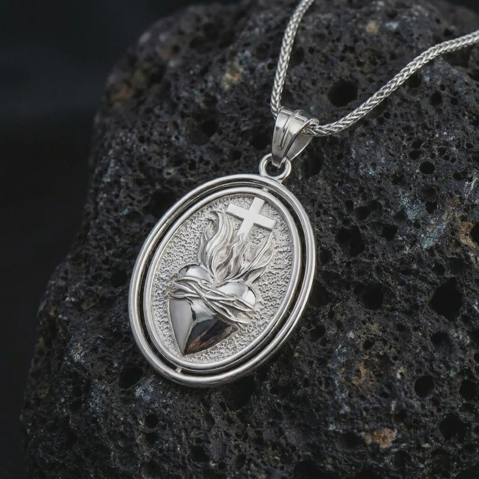 Polished silver Sacred Heart pendant necklace on textured volcanic rock, centered macro jewelry photo with detailed Christian relief and bright silver highlights.