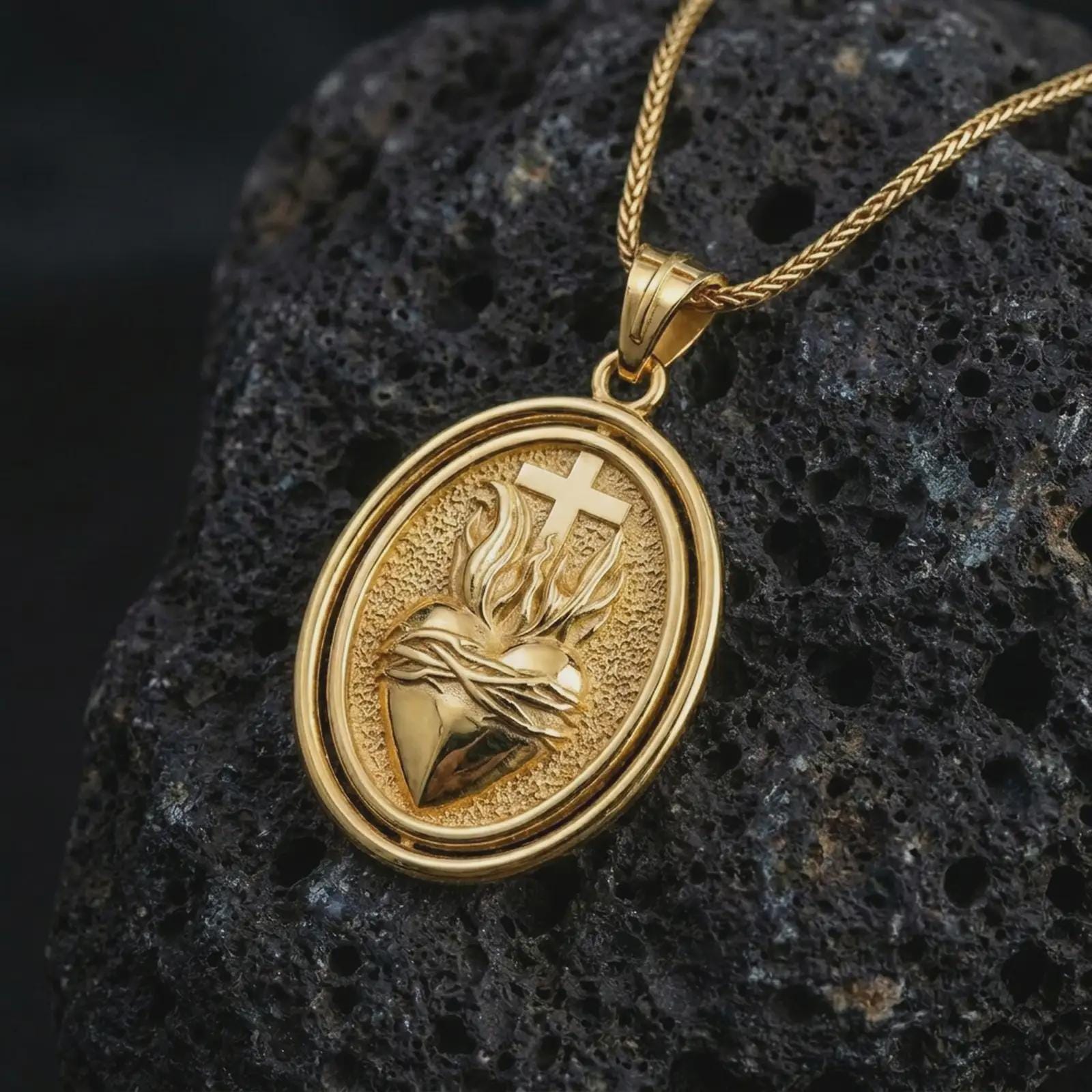 Gold plated Sacred Heart pendant necklace on textured volcanic rock, premium close-up studio composition emphasizing the flaming heart, cross, and crown of thorns symbolism.