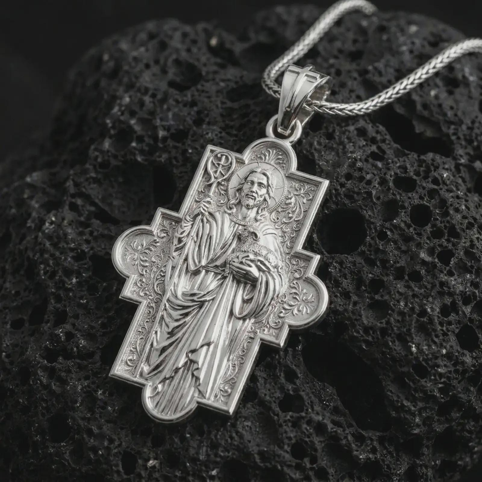 Polished silver Good Shepherd cross pendant necklace on textured volcanic rock, centered macro jewelry photo with detailed Christian icon relief and bright silver highlights.