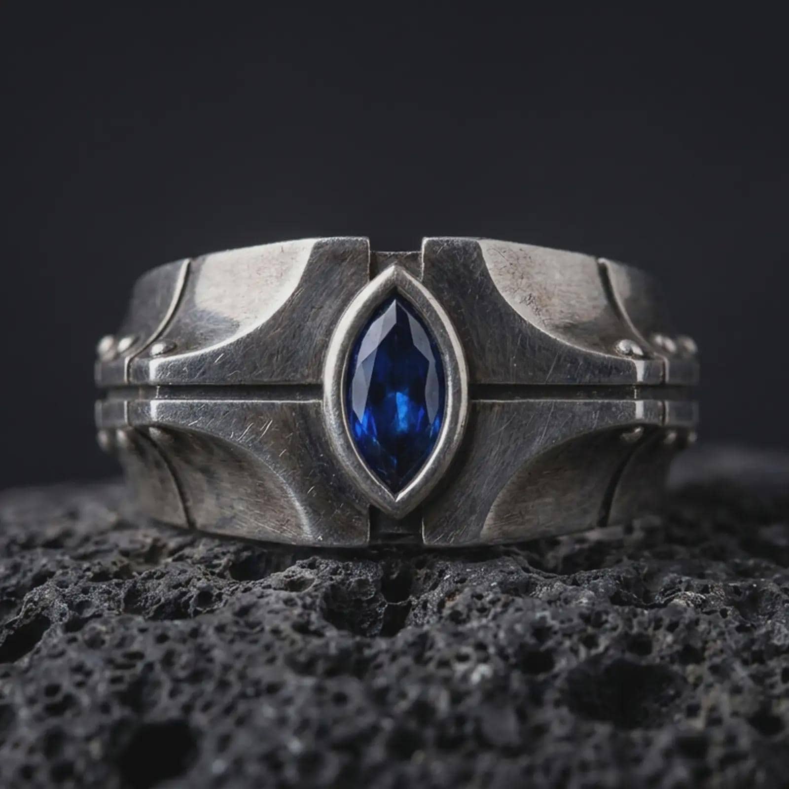 A sterling silver ring with an oxidized finish and marquise blue stone displayed on textured volcanic rock in a premium macro shot.