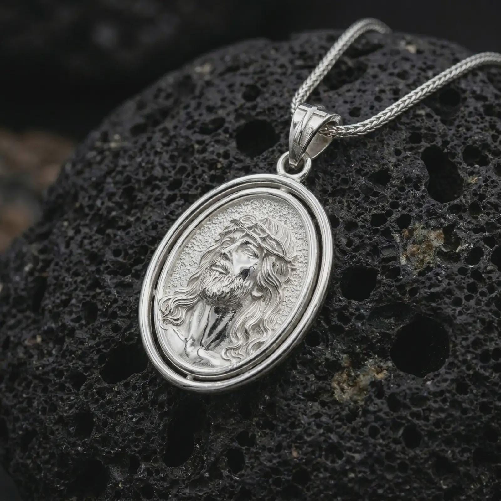 Polished silver spinning Jesus Christ pendant necklace on textured volcanic rock, centered macro jewelry photo with detailed Christian portrait relief and bright silver highlights.