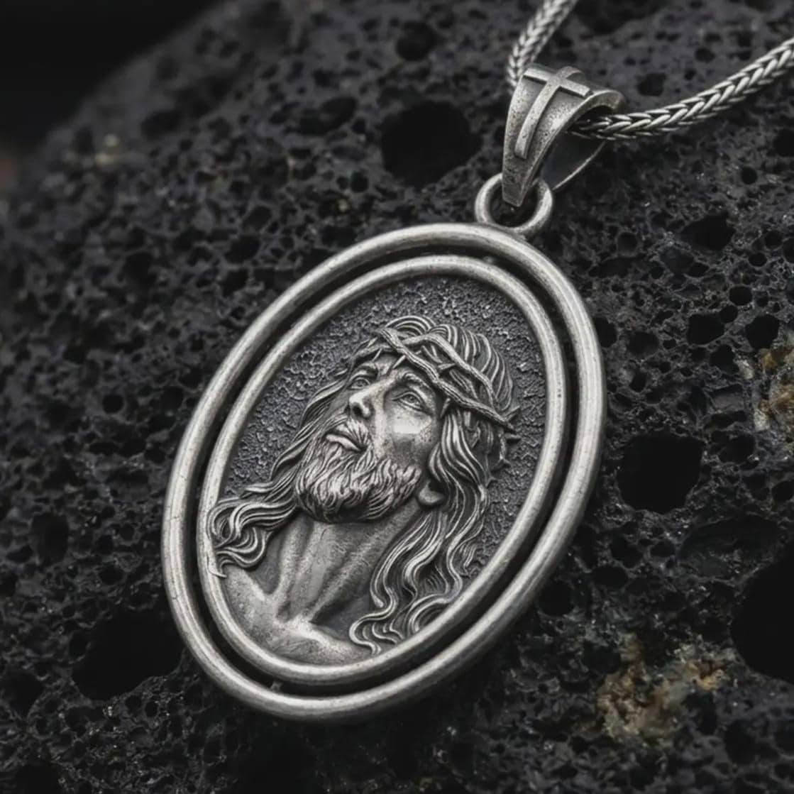 Oxidized spinning Jesus Christ pendant necklace on textured volcanic rock, close-up studio shot highlighting the crown of thorns portrait, oval frame, and antique silver contrast.