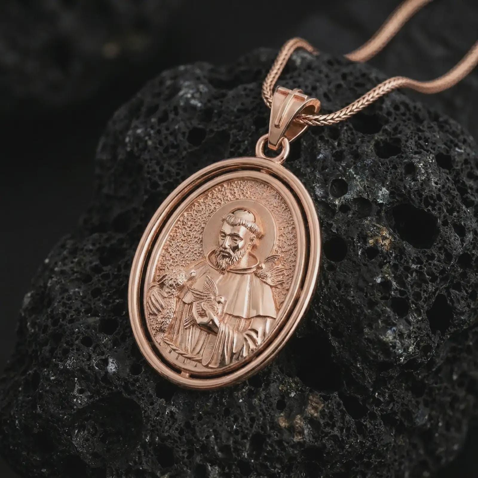 Rose gold plated Saint Francis pendant necklace on textured volcanic rock, detailed macro product shot with soft warm reflections and devotional Saint Francis design.