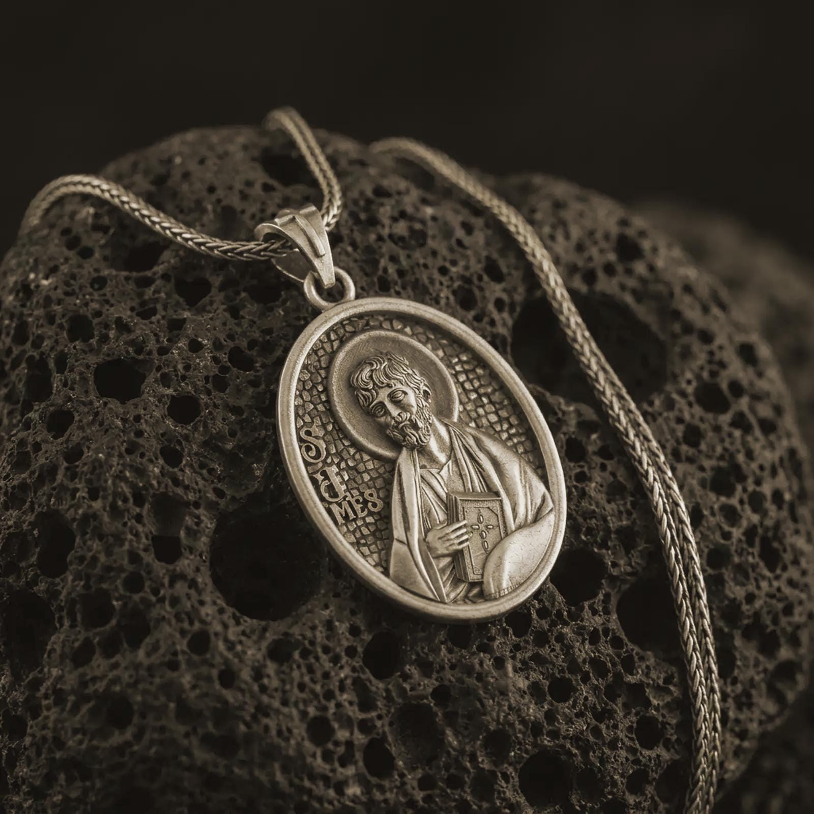 Bronze Saint James the Apostle pendant necklace on textured volcanic rock, close-up studio shot highlighting the apostle relief, halo, book detail, and antique silver contrast.