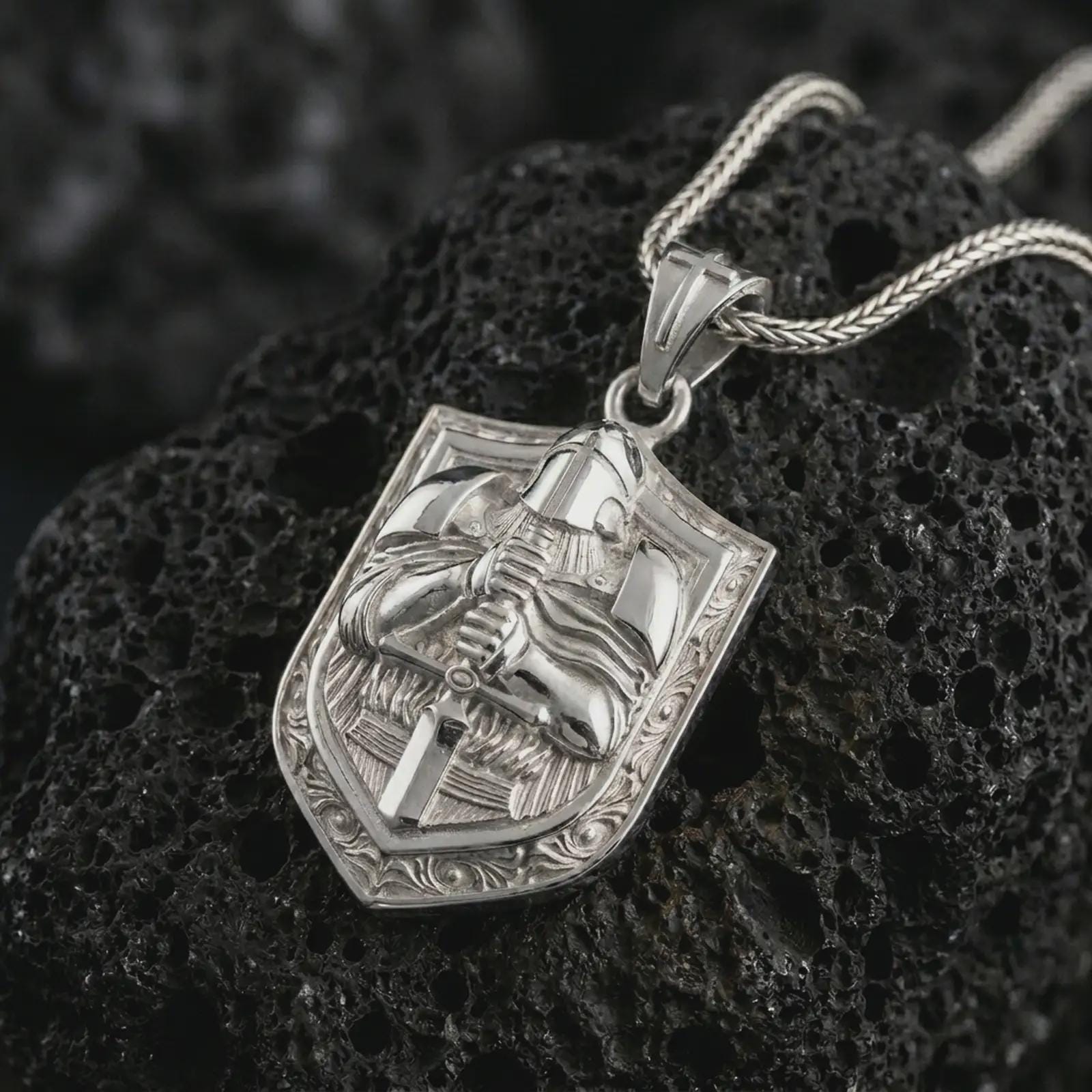 Polished silver knight shield pendant necklace on textured volcanic rock, centered macro jewelry photo with detailed medieval armor relief and bright silver highlights.