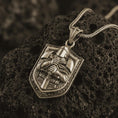 Load image into Gallery viewer, Bronze knight shield pendant necklace on textured volcanic rock, close-up studio shot highlighting the armored crusader figure, sword, shield shape, and antique silver contrast.

