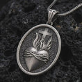 Load image into Gallery viewer, Oxidized Sacred Heart pendant necklace on textured volcanic rock, close-up studio shot highlighting the flaming heart, crown of thorns, cross, and antique silver contrast.
