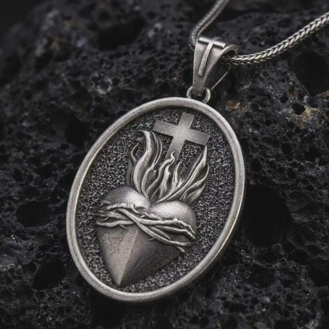 Oxidized Sacred Heart pendant necklace on textured volcanic rock, close-up studio shot highlighting the flaming heart, crown of thorns, cross, and antique silver contrast.