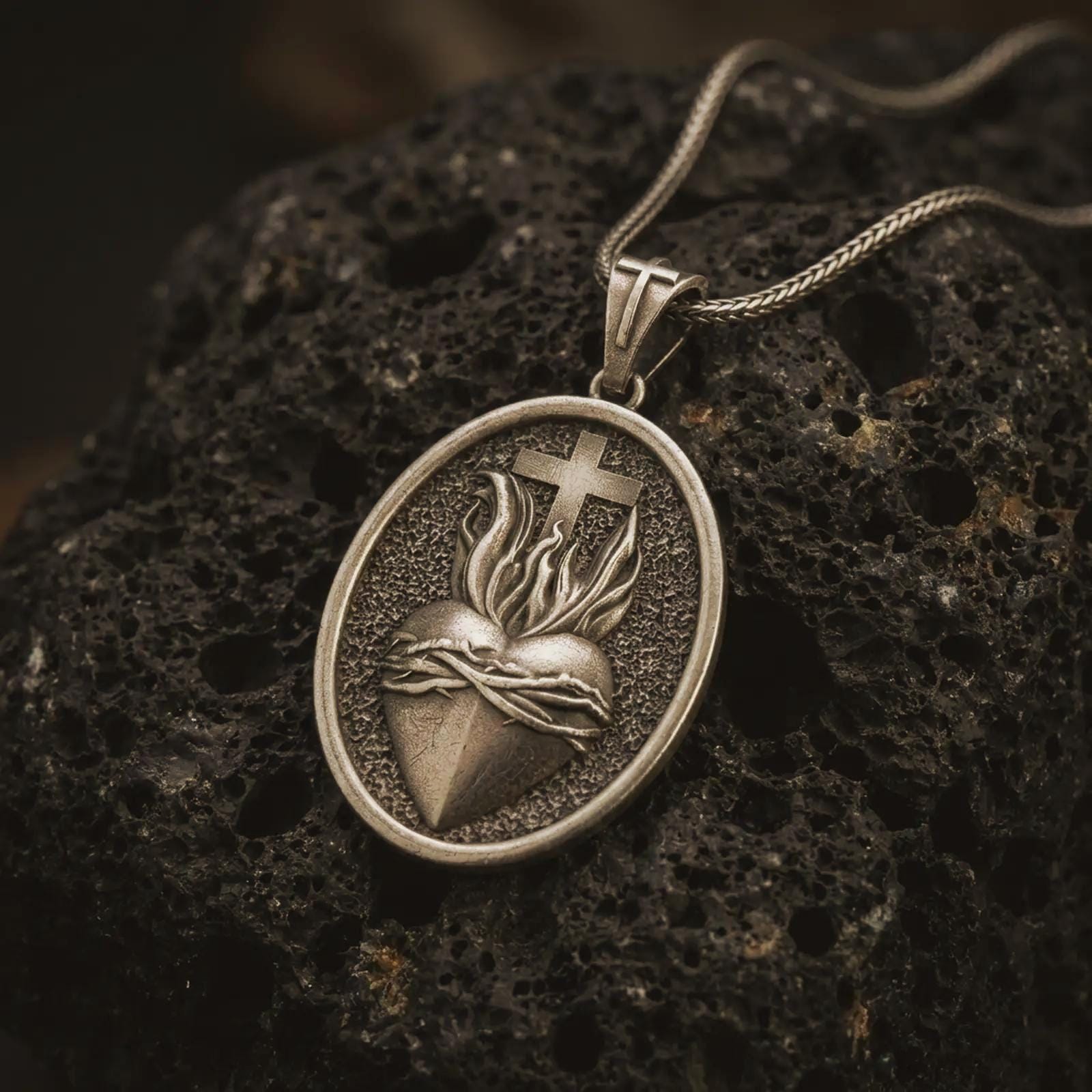 Bronze Sacred Heart pendant necklace on textured volcanic rock, close-up studio shot highlighting the flaming heart, crown of thorns, cross, and antique silver contrast.