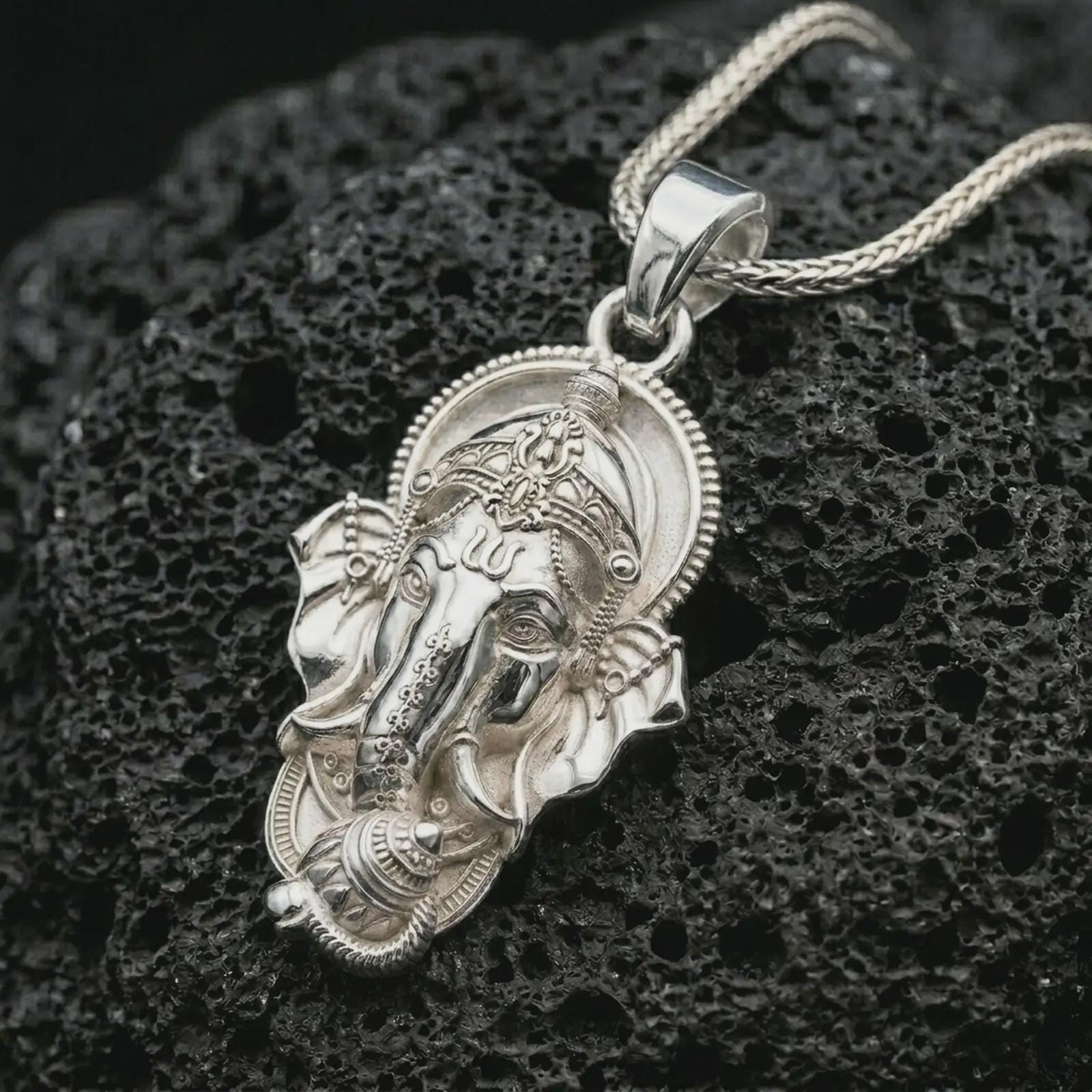 Polished silver Ganesha pendant necklace on textured volcanic rock, centered macro jewelry photo with detailed Hindu deity relief and bright silver highlights.