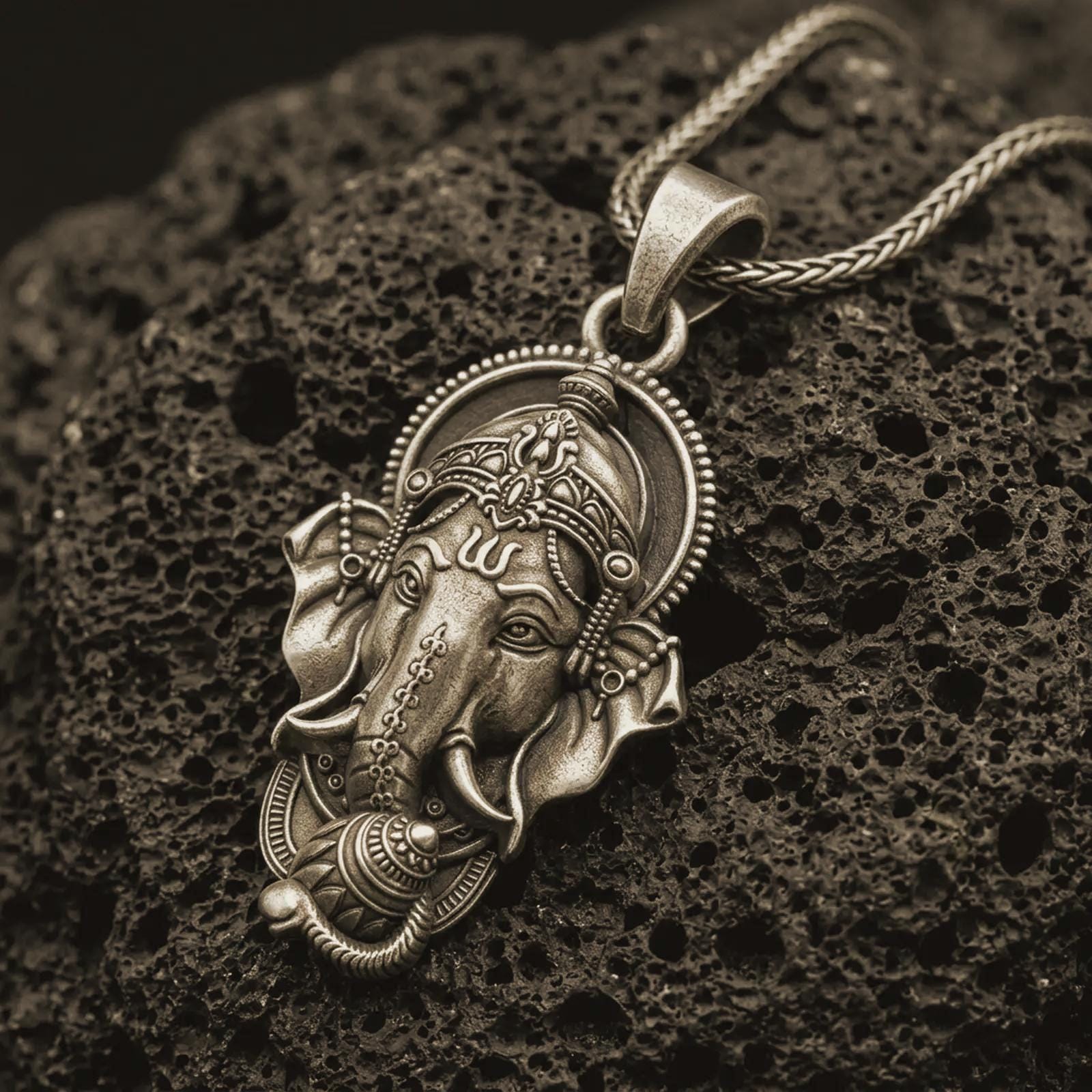 Bronze Ganesha pendant necklace on textured volcanic rock, close-up studio shot highlighting the elephant god face, ornate crown, tusks, and antique silver contrast.
