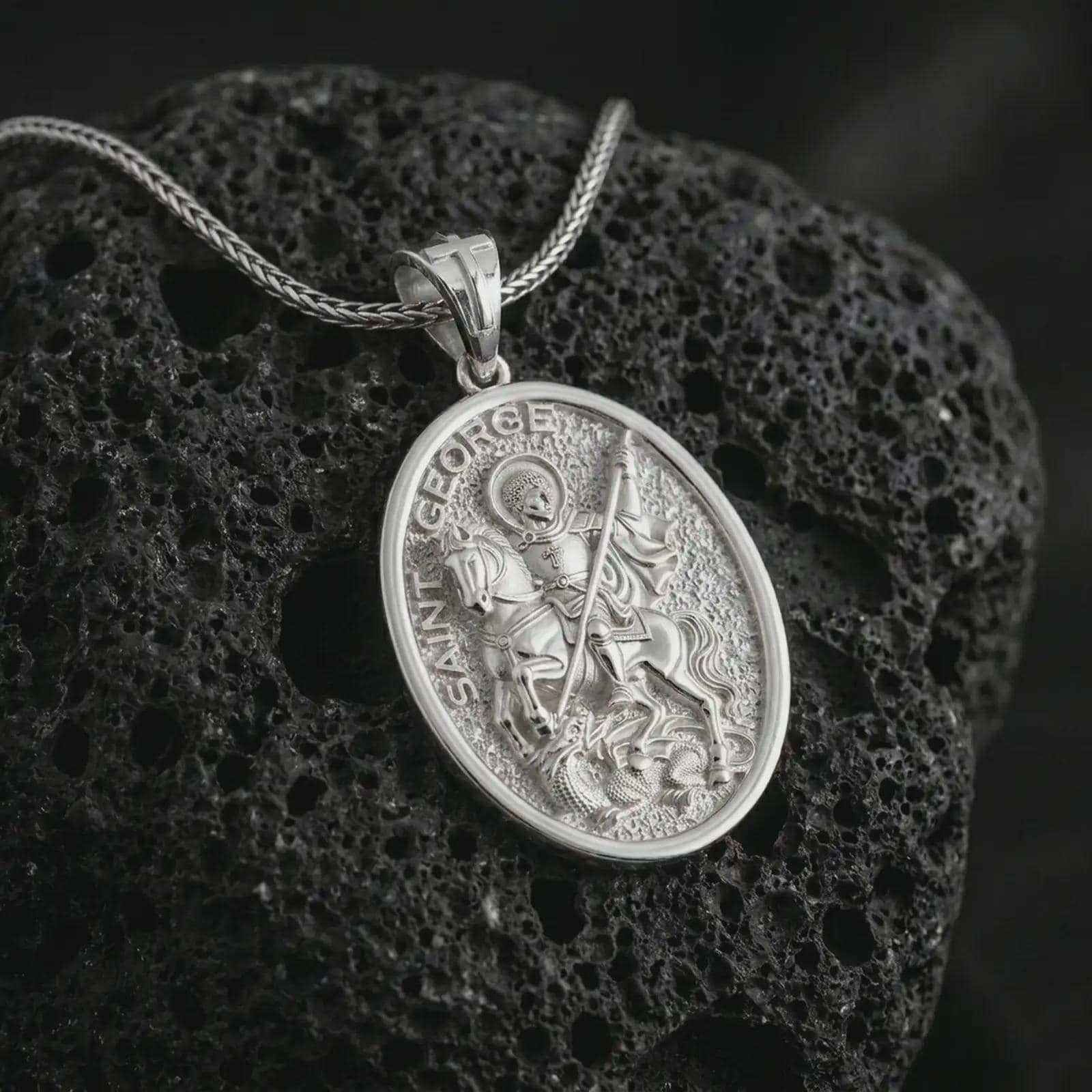 Polished silver Saint George pendant necklace on textured volcanic rock, centered macro jewelry photo with detailed dragon slayer relief and bright silver highlights.