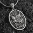 Load image into Gallery viewer, Oxidized Saint George pendant necklace on textured volcanic rock, close-up studio shot highlighting the mounted saint, spear, dragon detail, and antique silver contrast.
