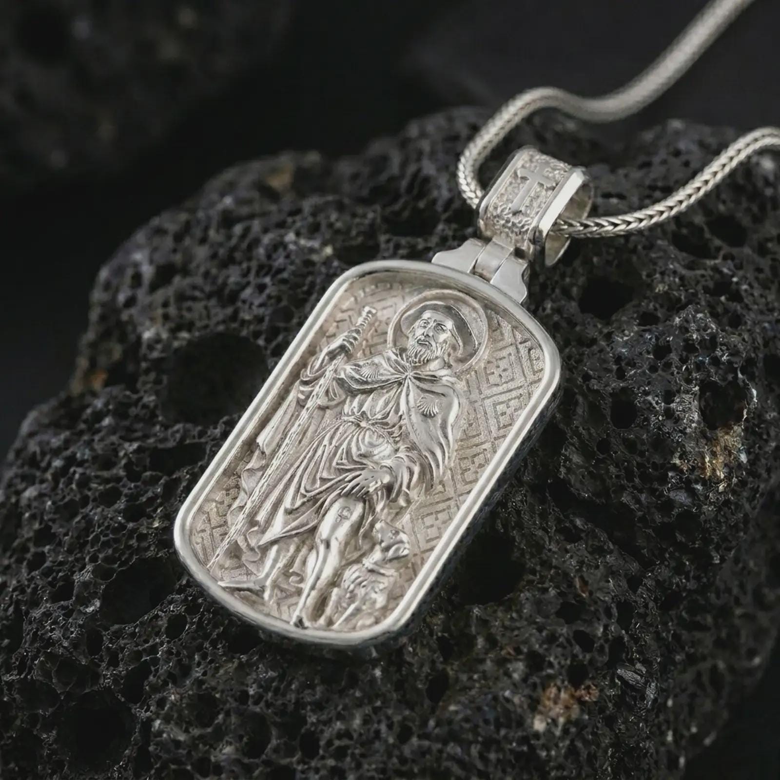 Polished silver Saint Roch dog tag pendant necklace on textured volcanic rock, centered macro jewelry photo with detailed Catholic patron medal engraving and bright silver highlights.