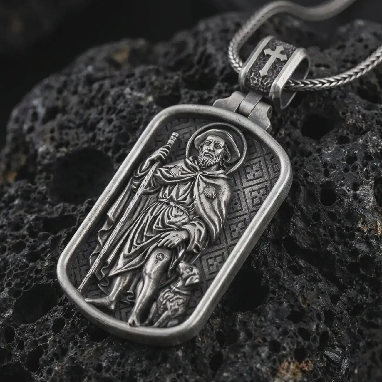 Oxidized Saint Roch dog tag pendant necklace on textured volcanic rock, close-up studio shot highlighting the pilgrim saint relief, staff, dog detail, and antique silver contrast.