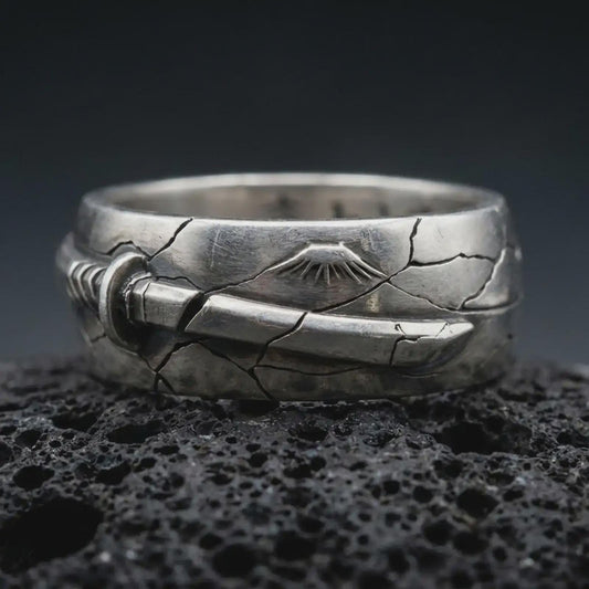 Oxidized Bushido sword band ring on textured volcanic rock, close-up studio shot highlighting the wrapped sword motif, cracked metal texture, rising sun detail, and antique silver contrast.