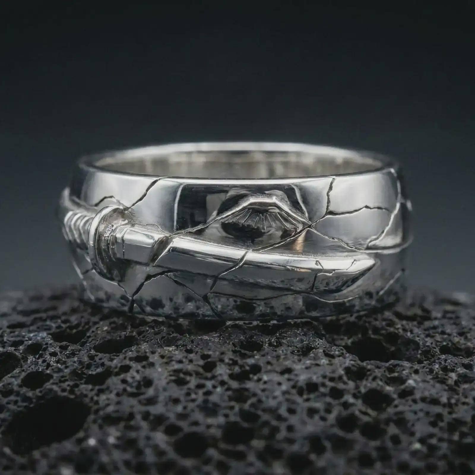 Polished silver Bushido sword band ring on textured volcanic rock, centered macro jewelry photo with detailed samurai-inspired sword relief and bright silver highlights.