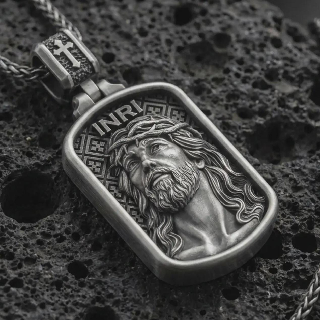 Oxidized INRI Jesus pendant necklace on textured volcanic rock, close-up studio shot highlighting the crown of thorns portrait, raised relief, and antique silver contrast.