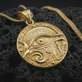 Load image into Gallery viewer, A gold plated Eye of Horus pendant lies on textured volcanic rock in a dark studio macro shot, highlighting the Egyptian protection amulet design.
