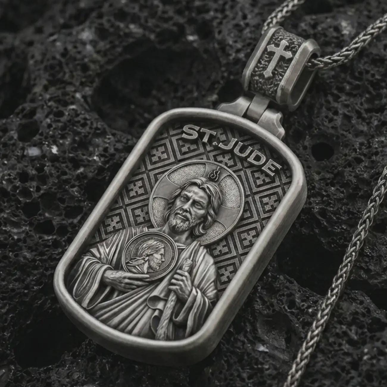 Oxidized Saint Jude dog tag pendant necklace on textured volcanic rock, close-up studio shot highlighting the saint relief, halo, medallion detail, and antique silver contrast.