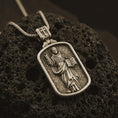 Load image into Gallery viewer, Bronze Saint Dominic dog tag pendant necklace on textured volcanic rock, close-up studio shot highlighting the saint relief, book, lily detail, and antique silver contrast.
