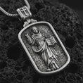 Load image into Gallery viewer, Oxidized Saint Dominic dog tag pendant necklace on textured volcanic rock, close-up studio shot highlighting the saint relief, book, lily detail, and antique silver contrast.
