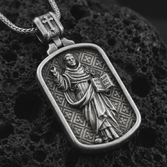 Oxidized Saint Dominic dog tag pendant necklace on textured volcanic rock, close-up studio shot highlighting the saint relief, book, lily detail, and antique silver contrast.