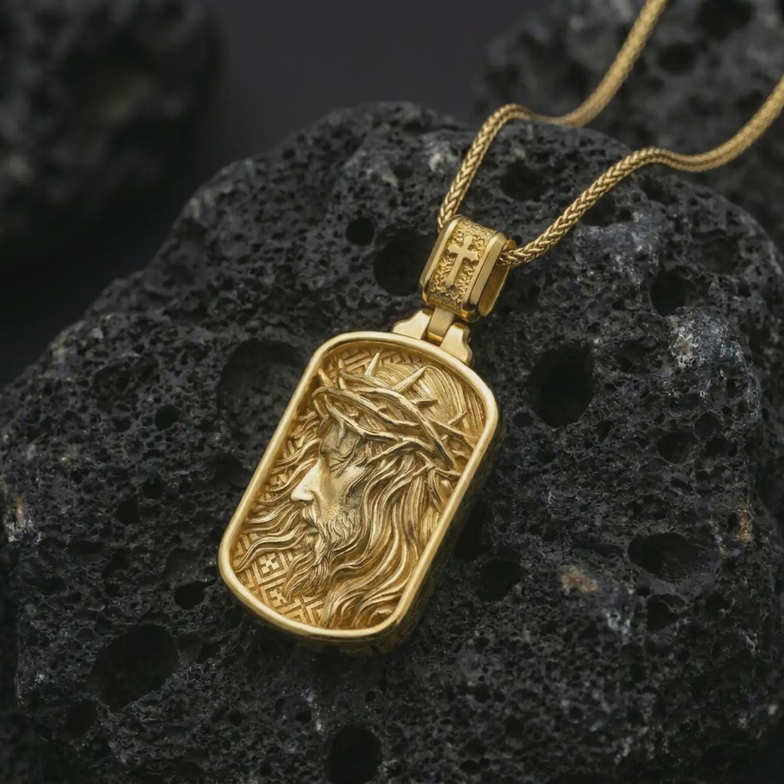 Gold plated crucified Jesus dog tag pendant necklace on textured volcanic rock, premium close-up studio composition emphasizing the side-profile Jesus figure, thorn crown, and rectangular medallion design.