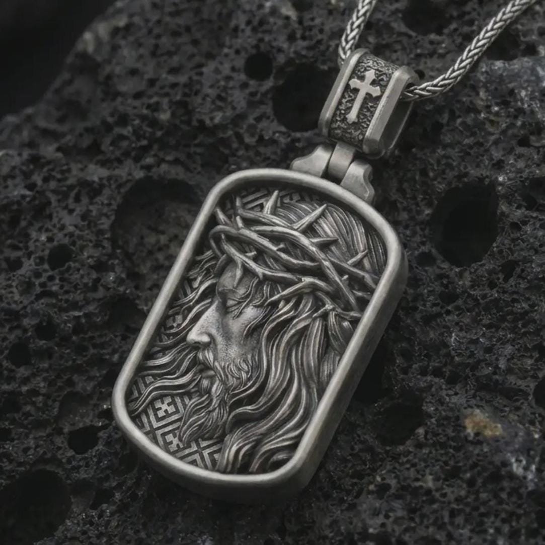 Oxidized crucified Jesus dog tag pendant necklace on textured volcanic rock, close-up studio shot highlighting the side-profile Christ relief, crown of thorns, flowing hair, and antique silver contrast.