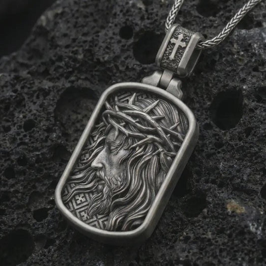 Oxidized crucified Jesus dog tag pendant necklace on textured volcanic rock, close-up studio shot highlighting the side-profile Christ relief, crown of thorns, flowing hair, and antique silver contrast.
