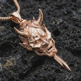 Load image into Gallery viewer, A rose gold plated oni mask pendant is photographed on porous volcanic rock in a close-up studio composition with bold luxury lighting.
