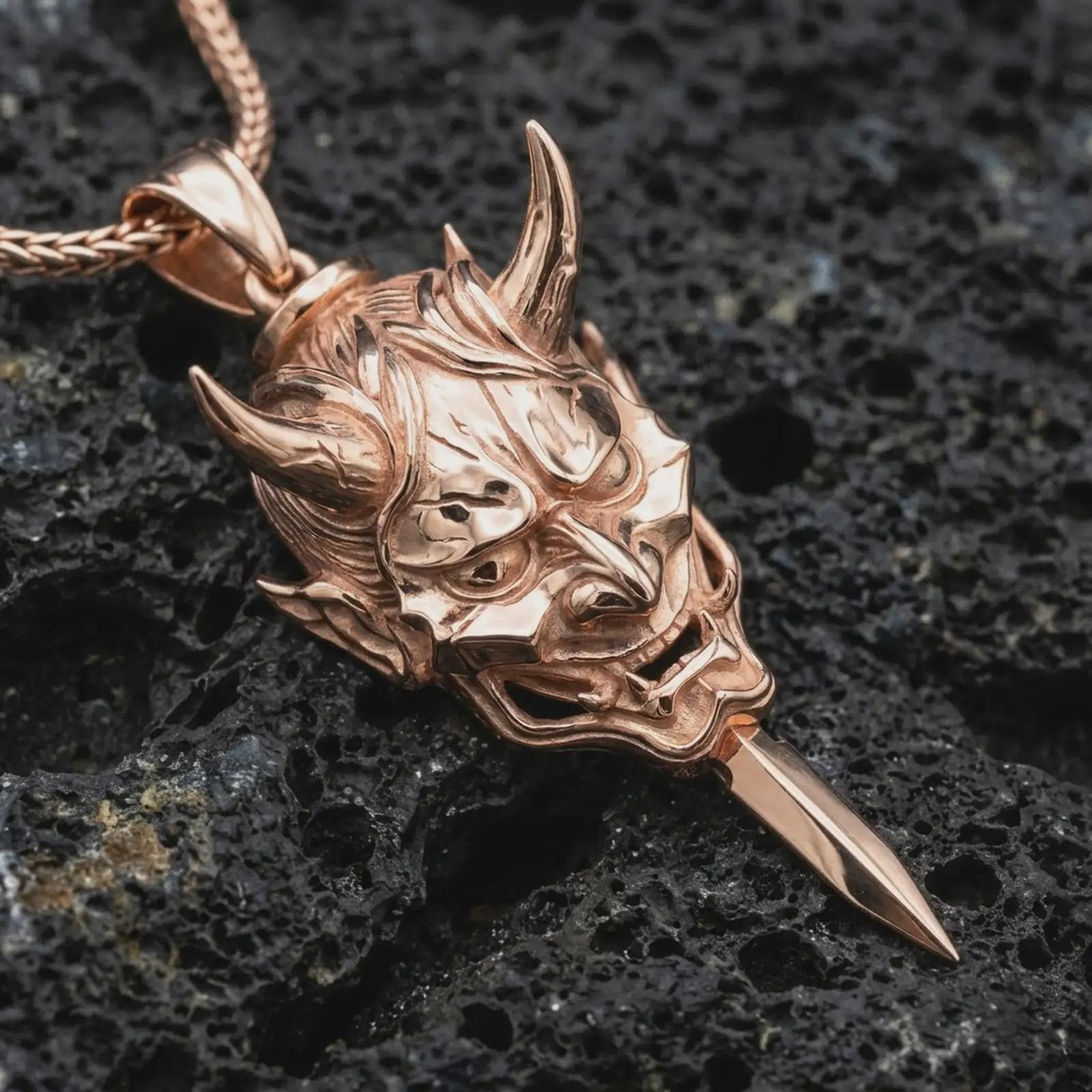A rose gold plated oni mask pendant is photographed on porous volcanic rock in a close-up studio composition with bold luxury lighting.