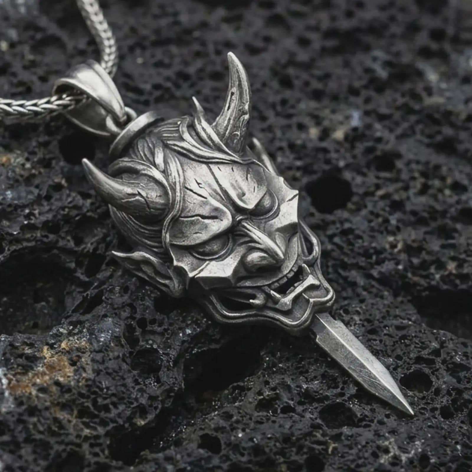An oxidized oni mask pendant rests on dark volcanic rock in a macro studio close-up, showing a fierce horned demon face with a blade-like detail.