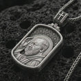 Load image into Gallery viewer, Oxidized Saint Padre Pio dog tag pendant necklace on textured volcanic rock, close-up studio shot highlighting the saint profile, blessing gesture, halo, and antique silver contrast.
