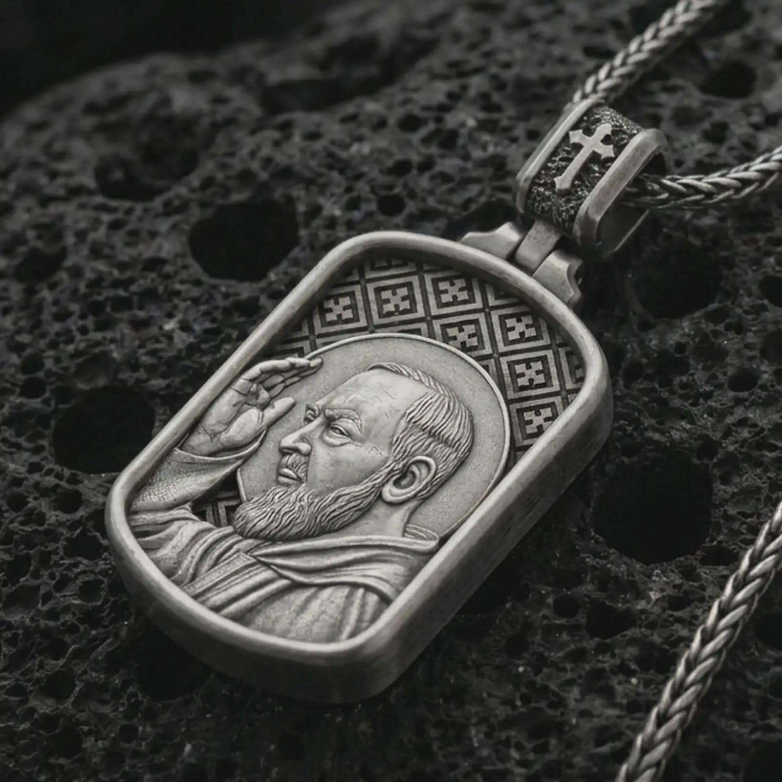 Oxidized Saint Padre Pio dog tag pendant necklace on textured volcanic rock, close-up studio shot highlighting the saint profile, blessing gesture, halo, and antique silver contrast.