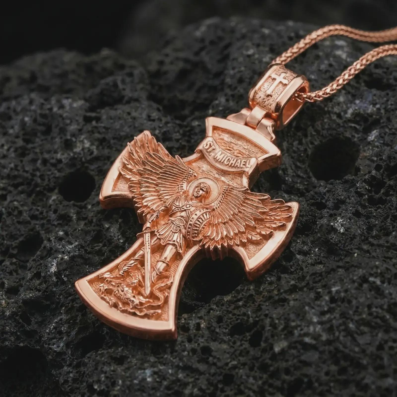 Rose gold plated Saint Michael cross pendant necklace on textured volcanic rock, detailed macro product shot with soft warm reflections and protective archangel design.