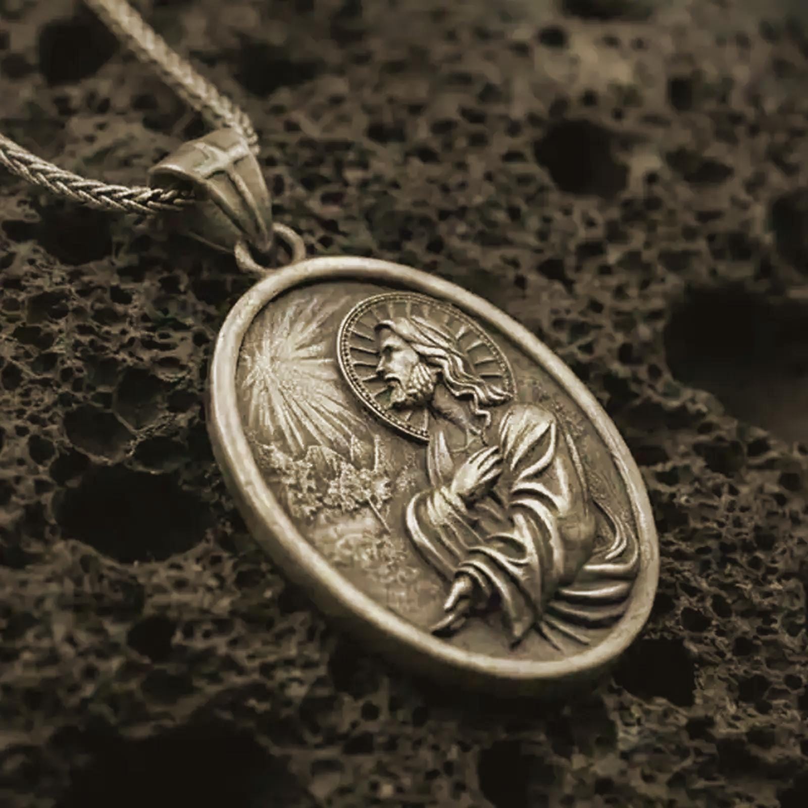 Bronze Jesus Christ prayer pendant necklace on textured volcanic rock, close-up studio shot highlighting the praying Christ relief, radiant light detail, halo, and antique silver contrast.
