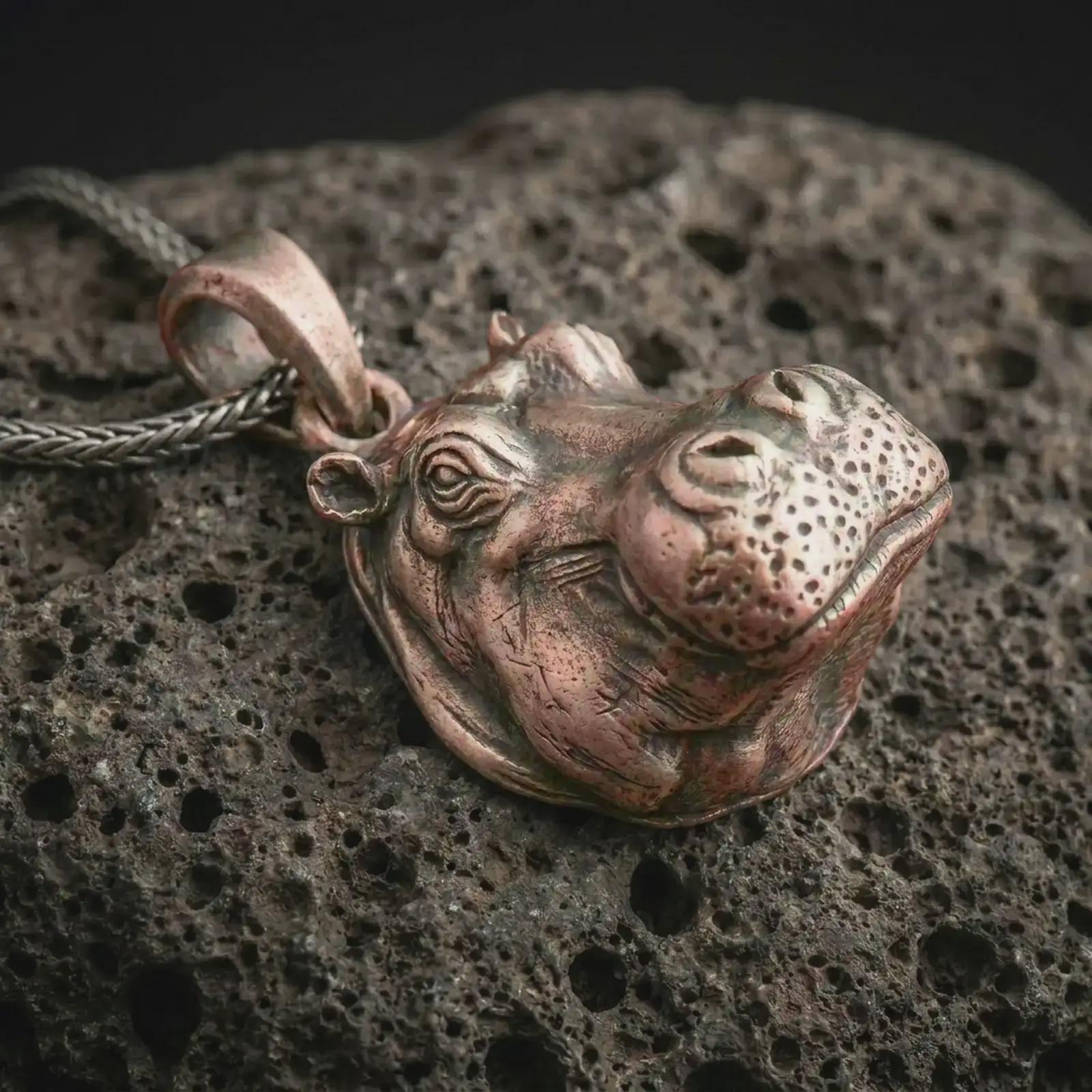 Bronze hippo head pendant necklace on textured volcanic rock, close-up studio shot highlighting the sculpted animal face, rounded muzzle, detailed eyes, and antique silver contrast.