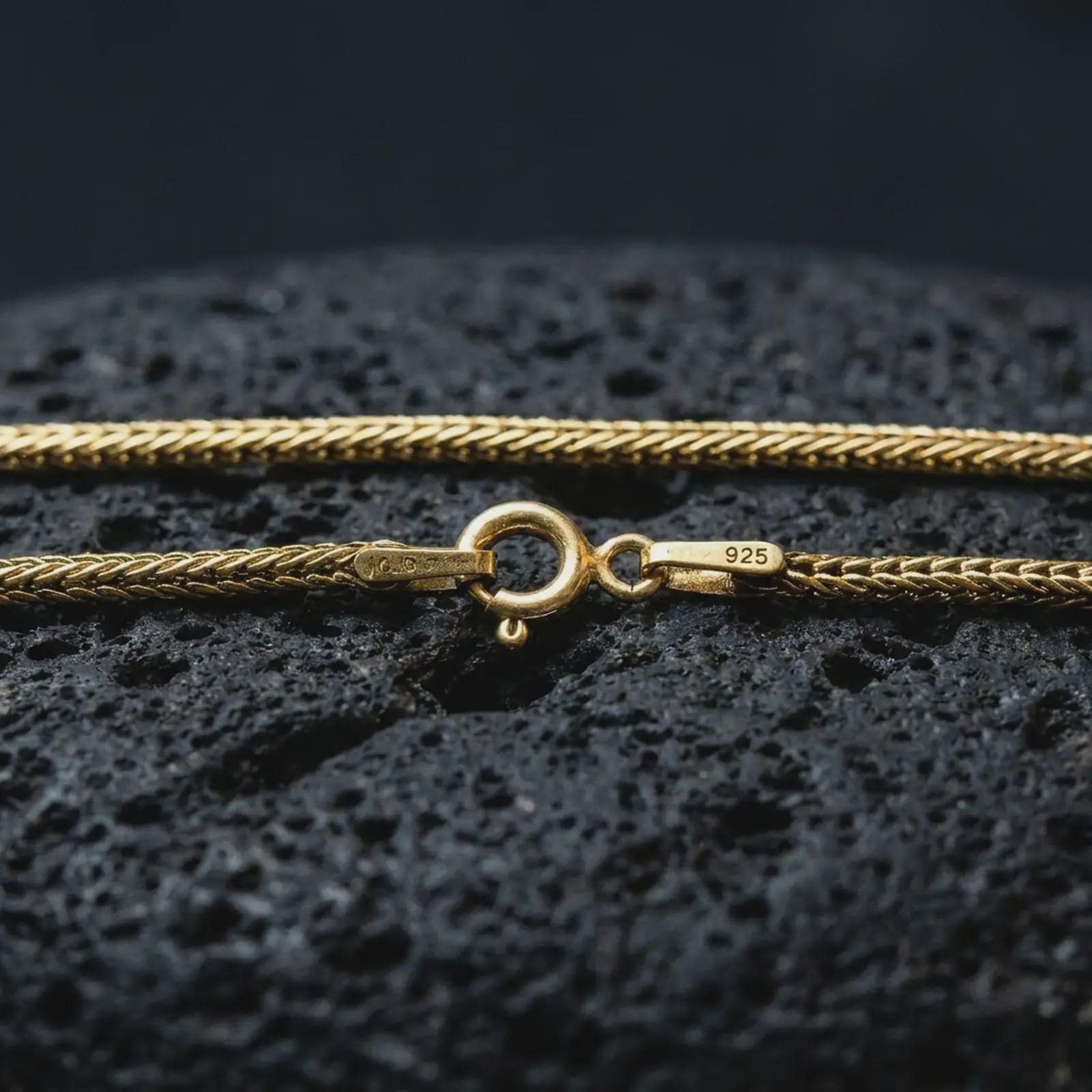 Gold plated Italian foxtail chain necklace on textured volcanic rock, premium close-up studio composition emphasizing the woven links, clasp detail, and refined chain craftsmanship.