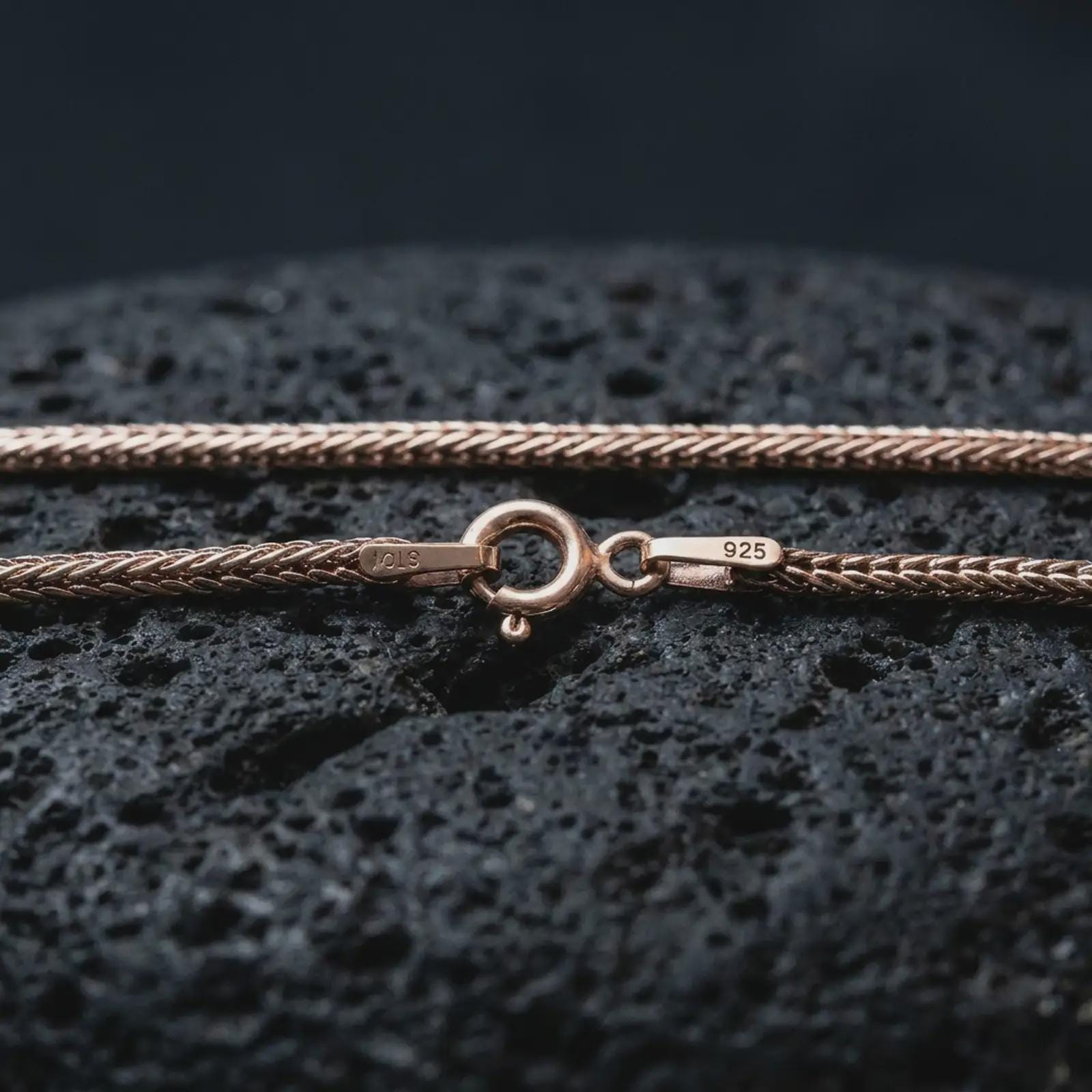 Rose gold plated Italian foxtail chain necklace on textured volcanic rock, detailed macro product shot with soft warm reflections and elegant spiga chain texture.
