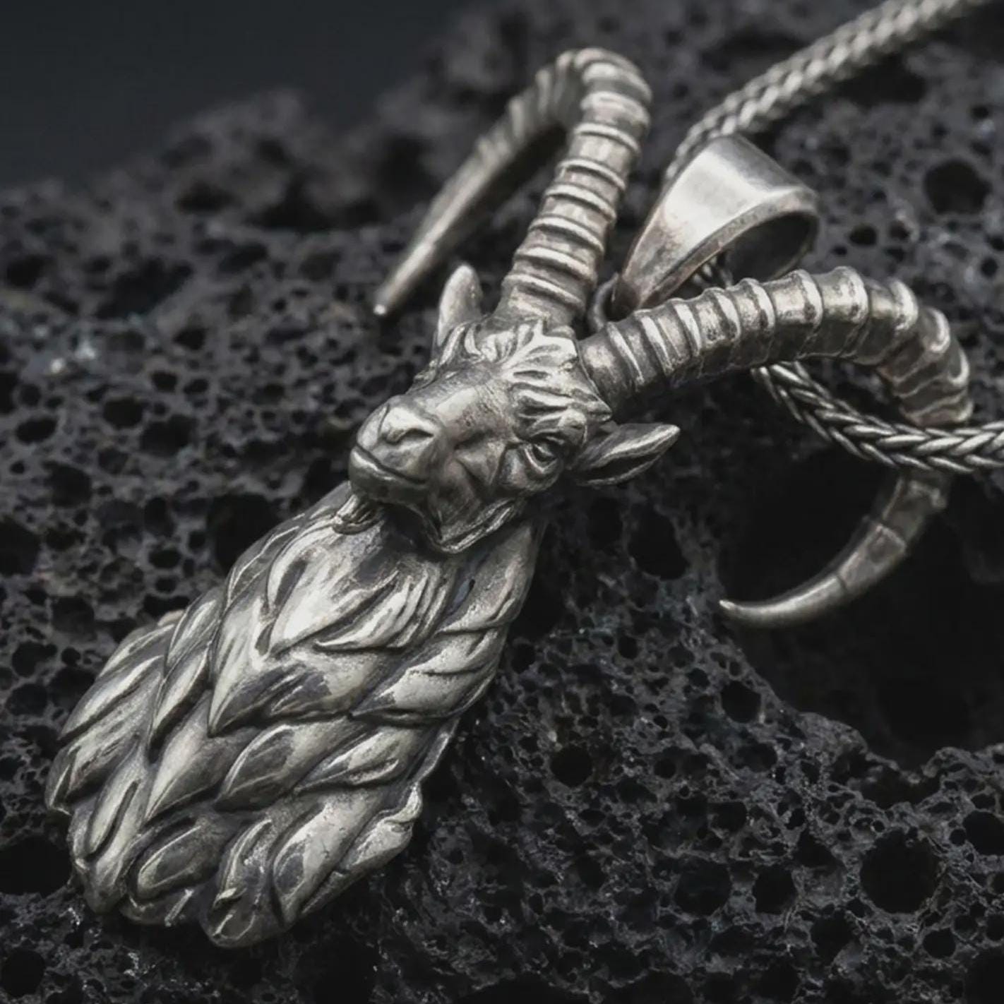 Oxidized ram horn pendant necklace on textured volcanic rock, close-up studio shot highlighting the sculpted goat head, curved horns, leaf-like beard detail, and antique silver contrast.
